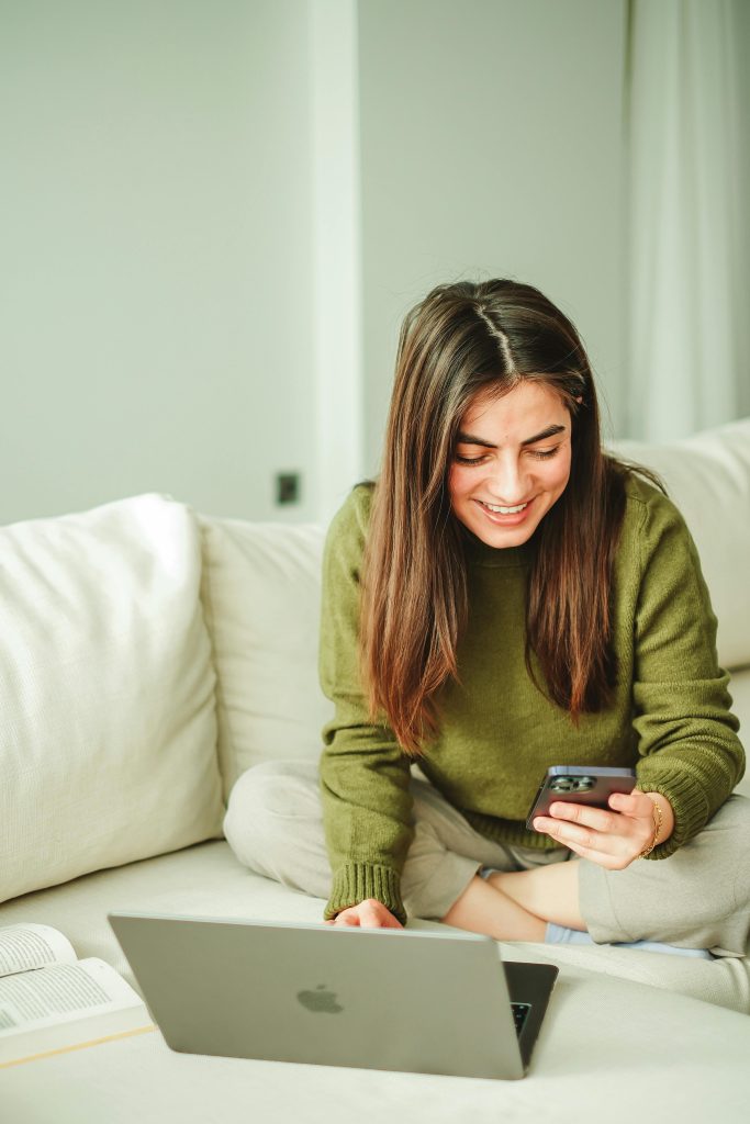 Young woman in cozy setting, engaging with smartphone and laptop, smiling while seated on sofa indoors.